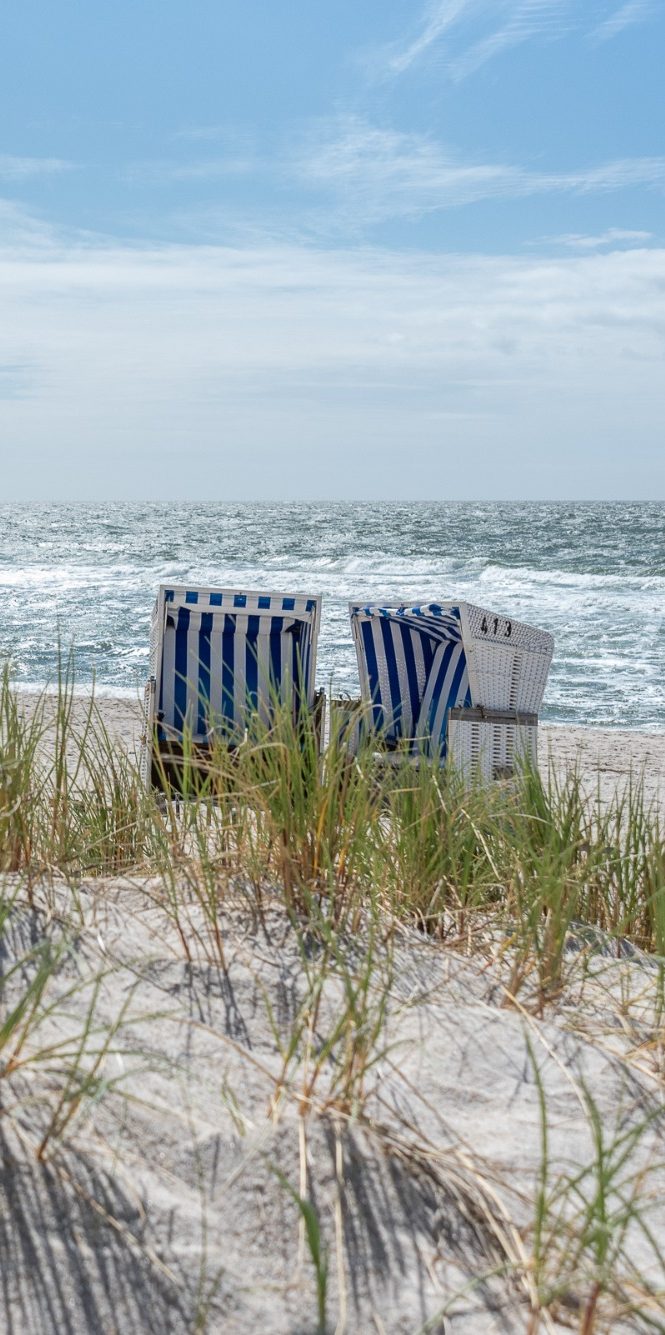 zwei blaugestreifte Strandkörbe stehen am Strand hinter Dünengras und Meer im Hintergrund